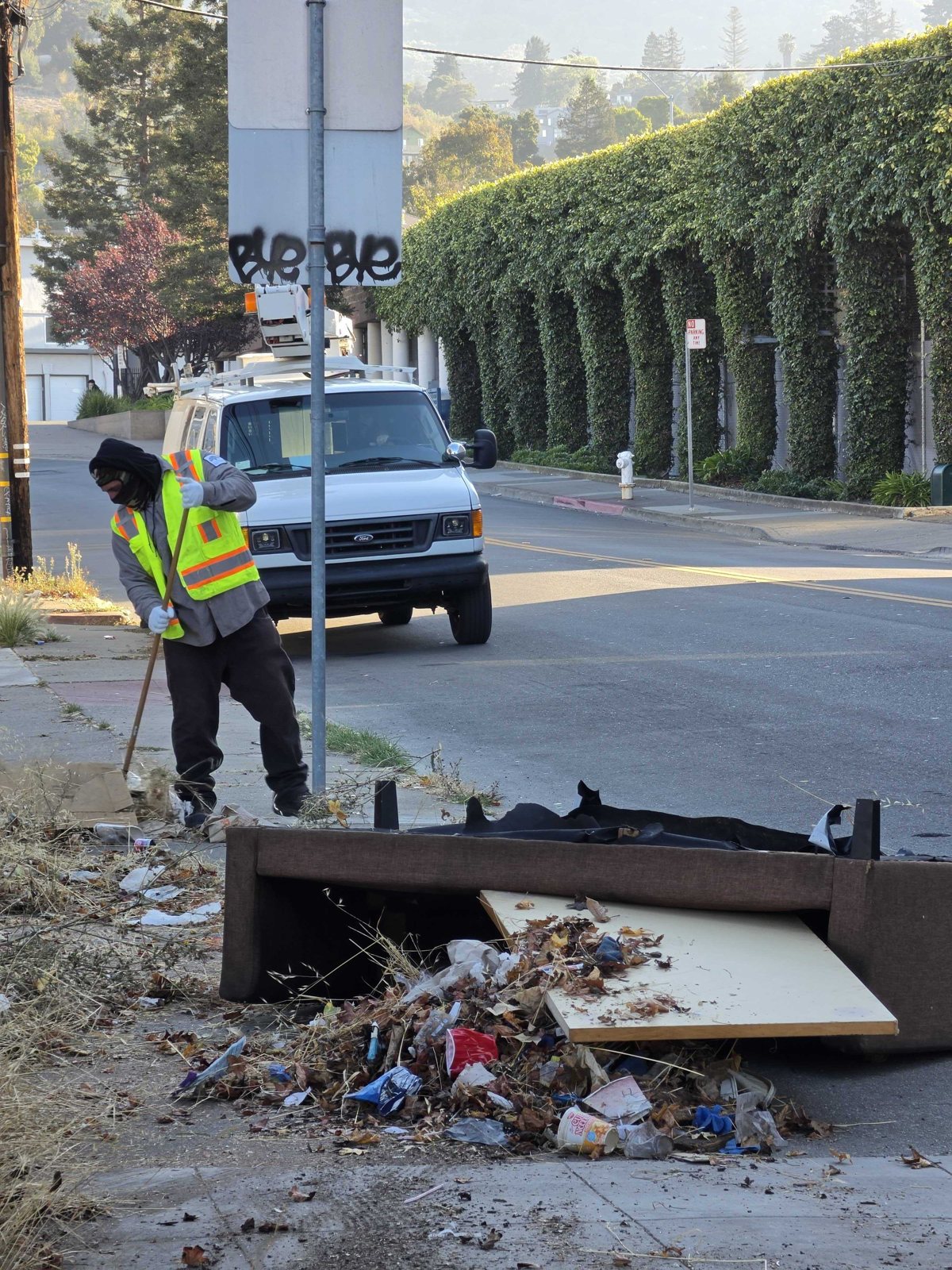 A crew member in a high-visibility vest raking debris beside a discarded mattress and box spring on a sidewalk
